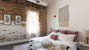 Ein Schlafzimmer mit Backsteinwand, einem Bett mit weißer und rosa Bettwäsche, einem Wandbehang aus Makramee und gerahmten Pflanzenbildern.