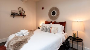 A neatly made bed with a fluffy throw, a bedside lamp, and a shelf with a book and a decorative item. - Thompson 212 · Thompson Block Loft - Modern and Historic (Ypsilanti)