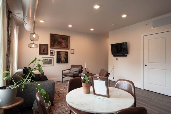 A modern living room with a round marble table, comfortable seating, a flat-screen TV mounted on the wall, and a large window with curtains. - Thompson 212 · Thompson Block Loft - Modern and Historic (Ypsilanti)