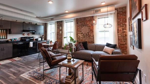 A modern living room with a sofa, chairs, a coffee table, and a kitchen in the background. - Thompson 212 · Thompson Block Loft - Modern and Historic (Ypsilanti)