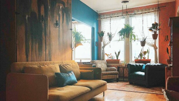 Sala de estar com sofá, poltronas e mesa de centro. Há plantas em vasos e uma janela com cortinas.