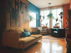 A living room with a sofa, armchairs, and a coffee table. There are potted plants and a window with curtains.