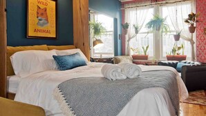 Quarto com cama, travesseiros, cobertor, quadro emoldurado na parede e plantas em vasos.