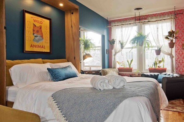 A bedroom with a bed, pillows, a blanket, a framed picture on the wall, and potted plants.