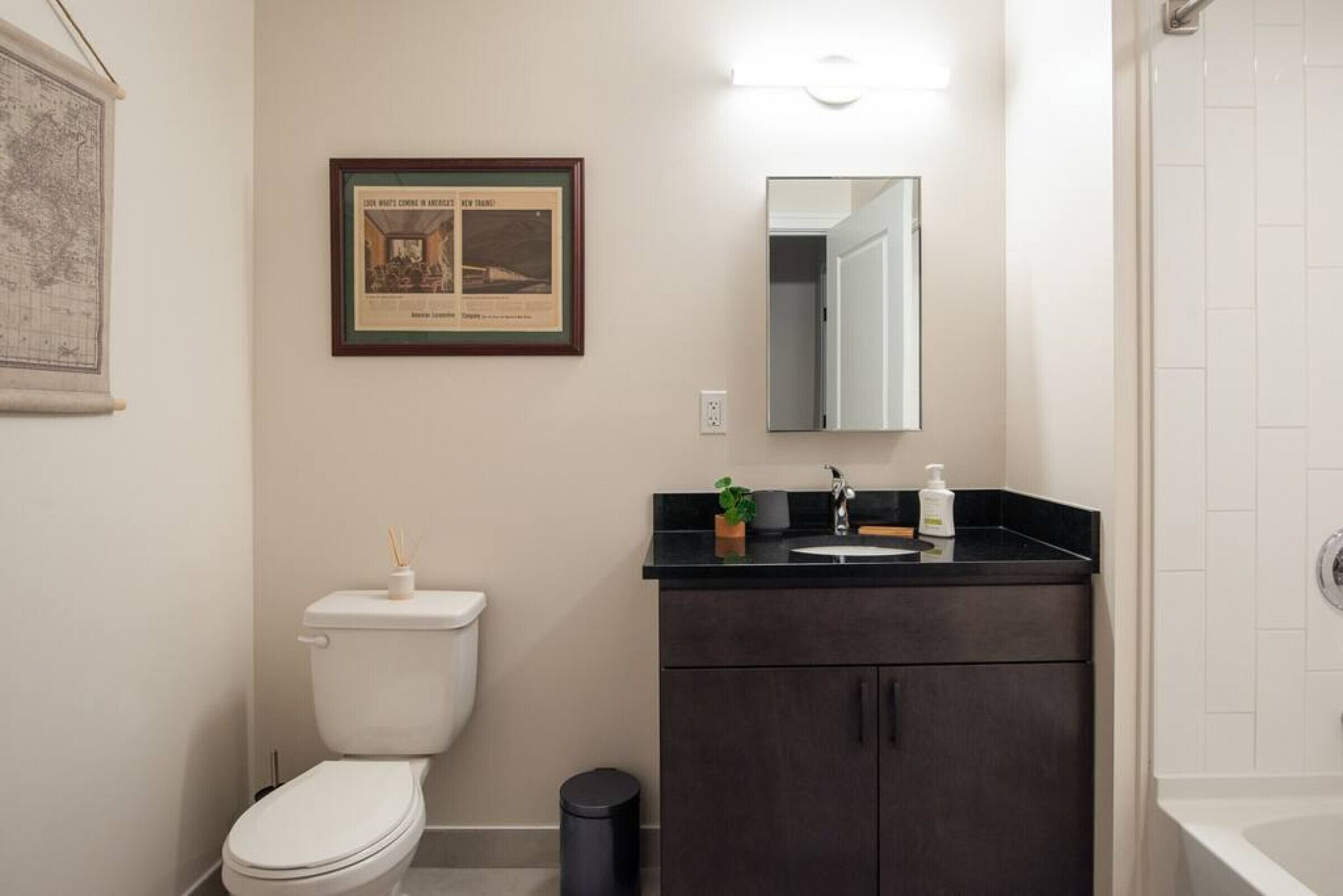 A bathroom with a toilet, sink, and mirror. There is a framed picture on the wall and a small trash can.