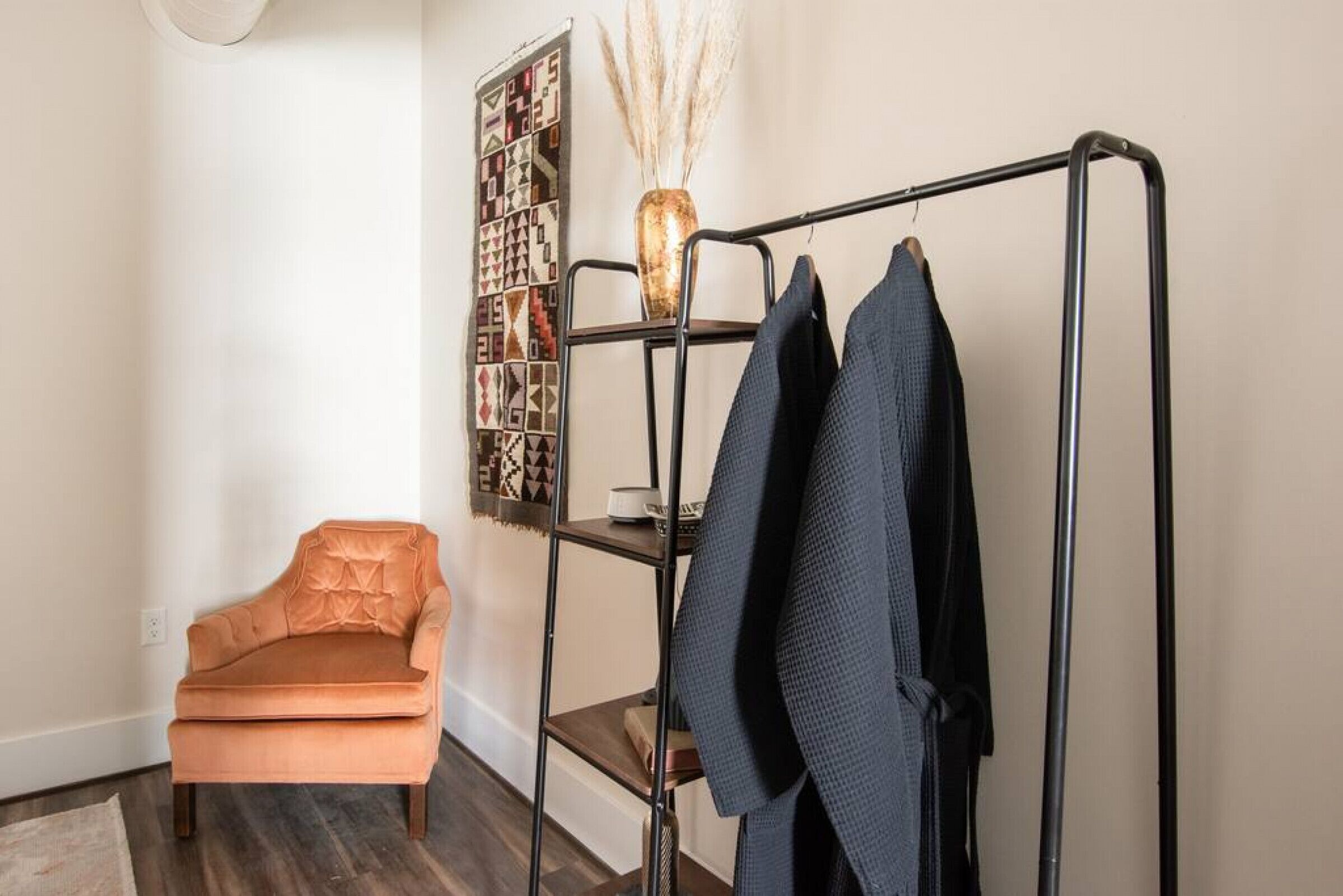 Een kamer met een zwart metalen rek met daarin een donker jack, een kleine oranje stoel, en een muur met kunstwerk met een geometrisch patroon.
