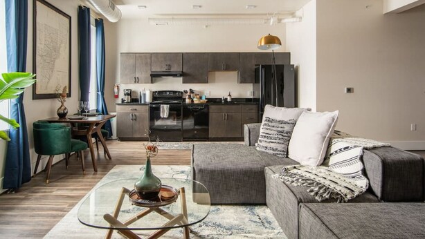 Una sala moderna con un sofá gris, una mesa de centro de vidrio y una zona de comedor con una silla verde. Se observa una cocina con armarios oscuros y una lámpara colgante al fondo.