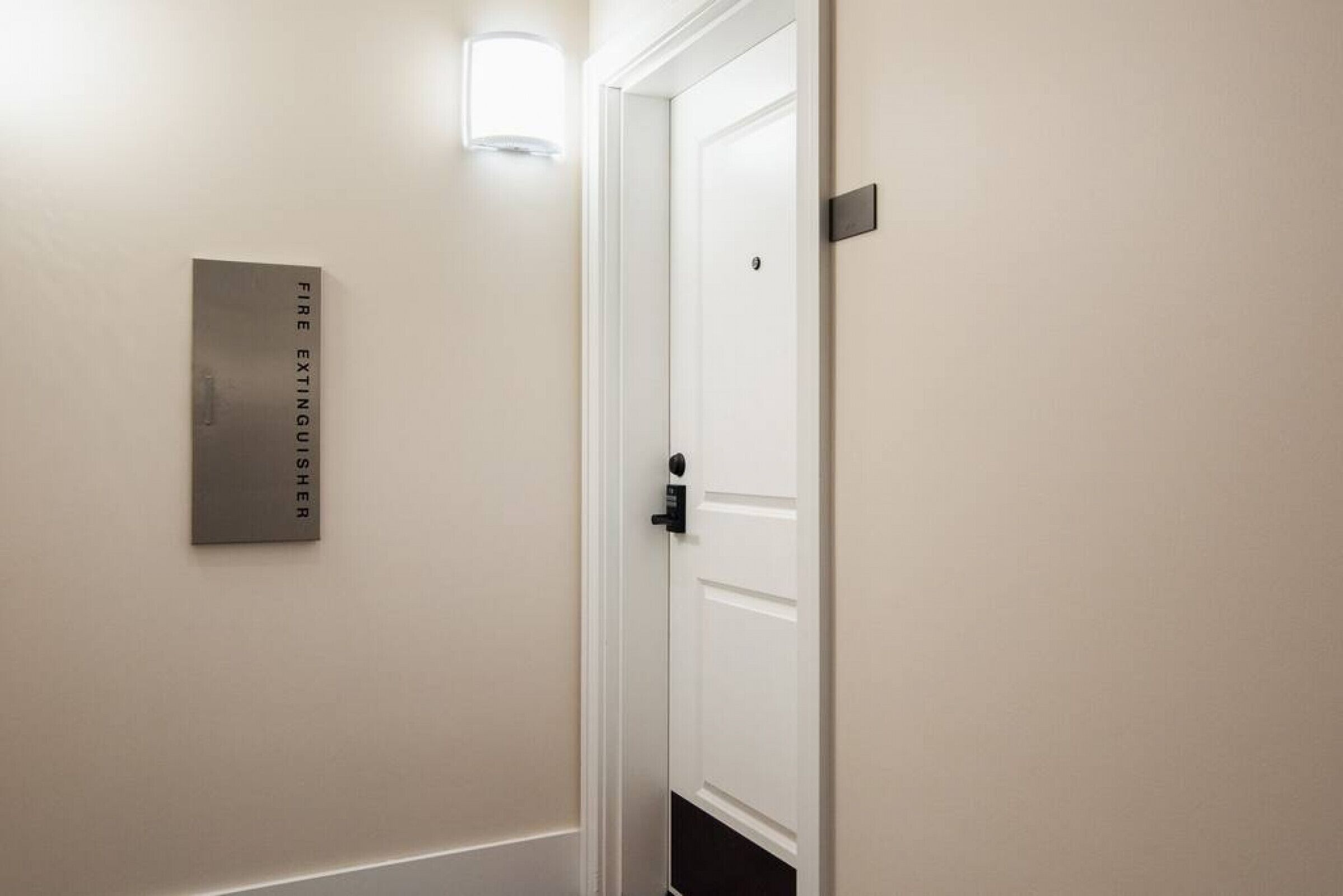 Un pasillo con una puerta blanca, un extintor montado en la pared y una lámpara sobre la puerta.