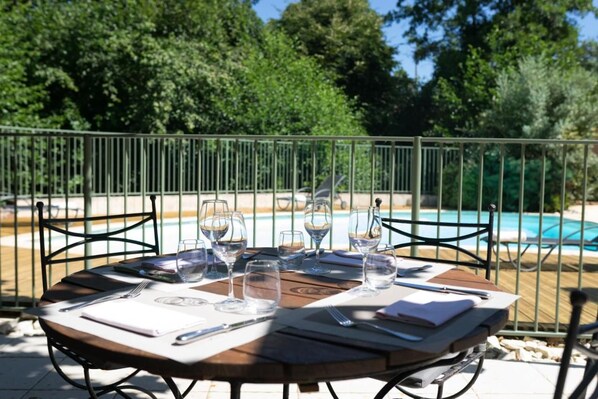 A round wooden table set for dining with a view of a swimming pool and trees in the background.