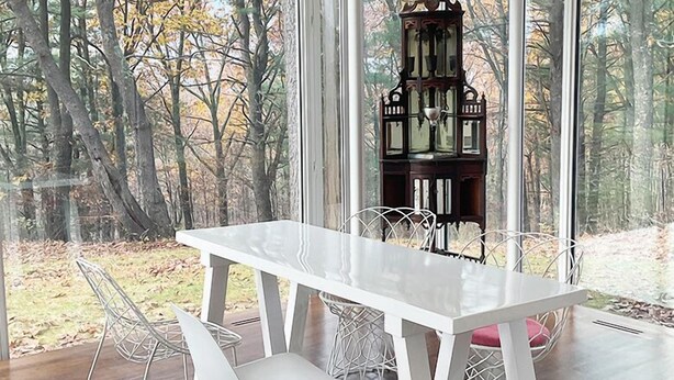 A dining area with a white table and chairs, a decorative lantern, and a view of a forest through large windows. - Modern Glass House designed by renowned architect Thomas Phifer (Hudson)