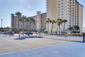 A tennis court with a net, surrounded by parked cars and palm trees.
