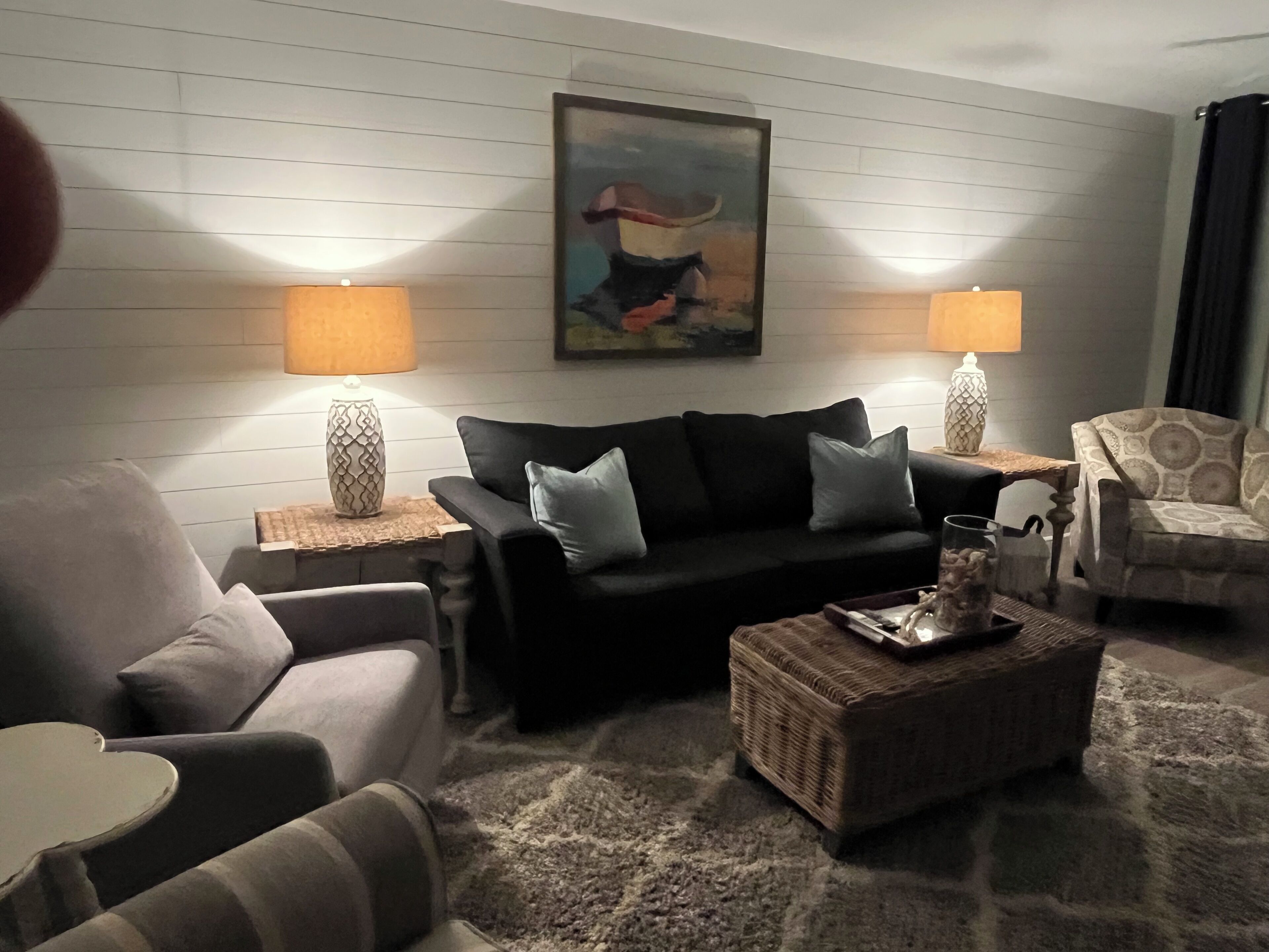 Un salon avec un canapé noir, deux tables d’appoint avec des lampes, un tapis à motifs et un tableau accroché au mur.