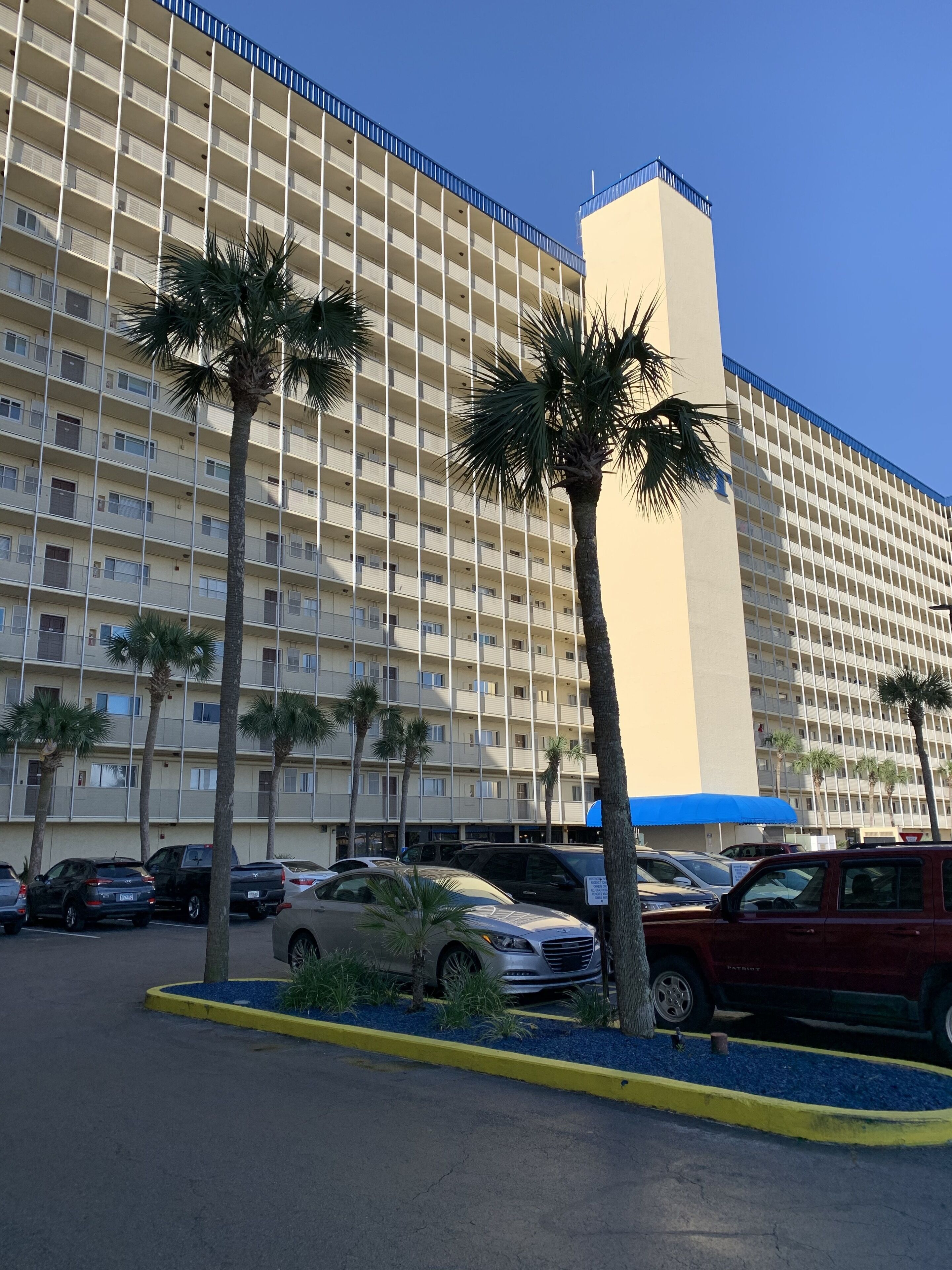 Un hotel de varios pisos con fachada color beige y detalles azules, que cuenta con un estacionamiento con varios automóviles y palmeras.