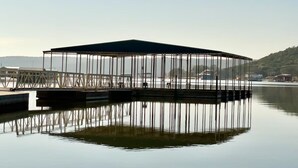 Eine hölzerne Stegkonstruktion mit Metallgestell und Dach, auf ruhigem Wasser mit spiegelnder Oberfläche.