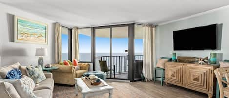 A spacious living room with a large window overlooking the sea, a comfortable sofa set, a wooden cabinet, and a flat-screen TV mounted on the wall.