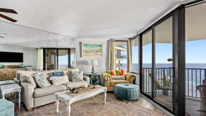 A spacious living room with a large window offering a view of the ocean, a beige sofa, a white coffee table, and a small side table with a lamp. - Sunbird 1103 West (Panama City Beach)