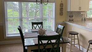 A dining area with a table, chairs, and a chandelier. There is a window with blinds, a kitchen area with cabinets, and a countertop.