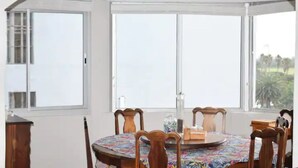 Un coin repas avec une table ronde, des chaises en bois et une nappe colorée. De grandes fenêtres offrent une vue sur l’extérieur.