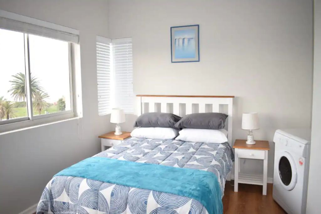A bedroom with a bed, two bedside tables with lamps, a washing machine, a window with blinds, and a painting on the wall.