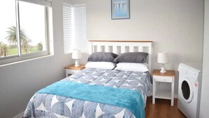 Une chambre à coucher avec un lit, deux tables de chevet avec des lampes, un lave-linge, une fenêtre avec des stores et un tableau accroché au mur.