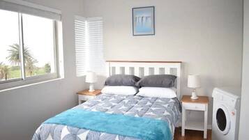 A bedroom with a bed, two bedside tables with lamps, a washing machine, a window with blinds, and a painting on the wall.