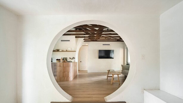 Vista interior a través de un arco que da a una sala de estar con suelo de madera, un sofá y un televisor montado en la pared.