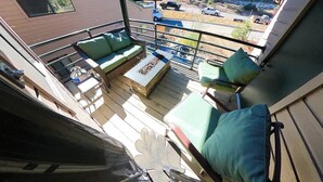A balcony with a wooden deck, green cushioned seating, and a view of a residential area with buildings and parked cars.