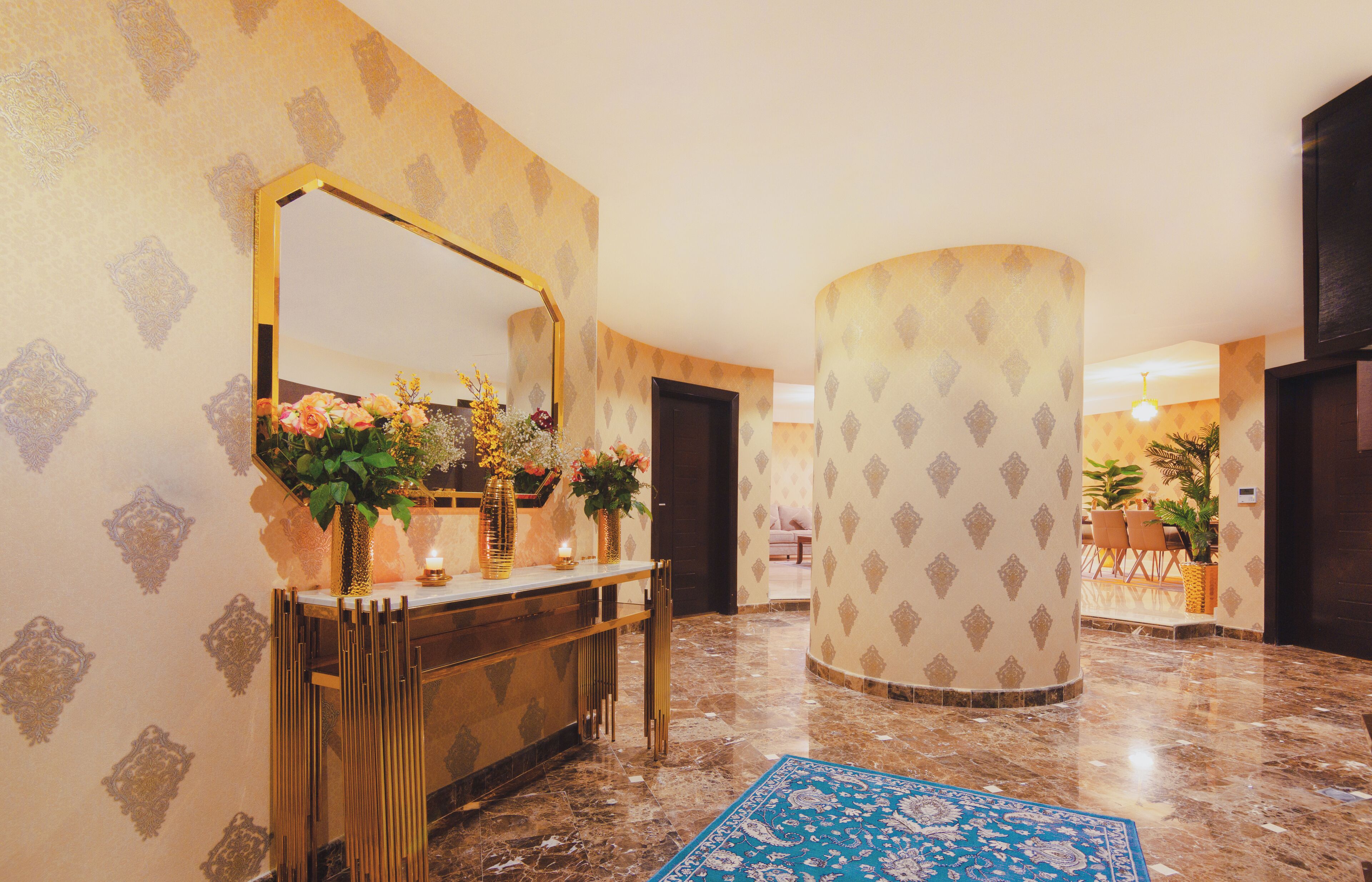 A lobby with a marble floor, a gold-framed mirror, a console table with floral arrangements, and patterned wallpaper.
