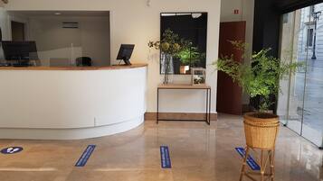 Un comptoir d’accueil avec un ordinateur, une horloge et un vase rempli de fleurs. On y trouve également une plante en pot et un panneau posé sur le sol.
