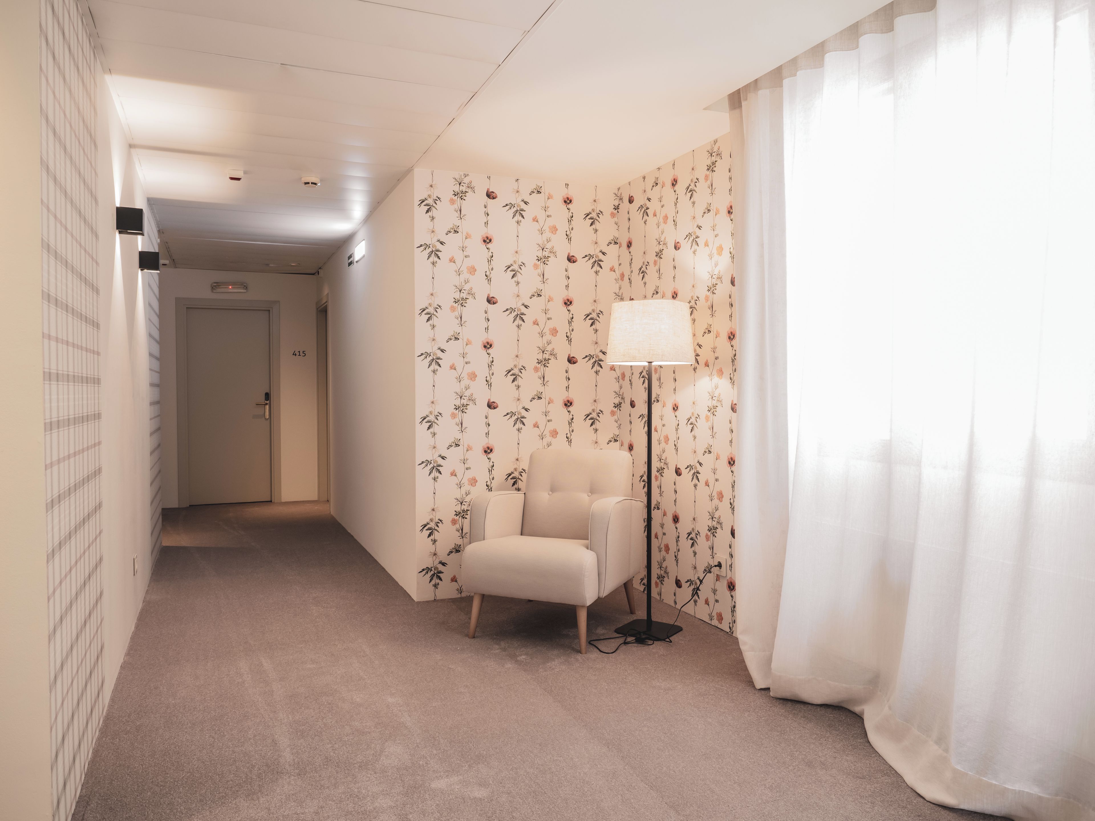 A hotel room with a floral wallpaper, a white armchair, a floor lamp, and a view of a hallway with a door marked '415'.