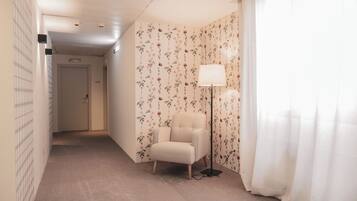 A hotel room with a floral wallpaper, a white armchair, a floor lamp, and a view of a hallway with a door marked '415'.