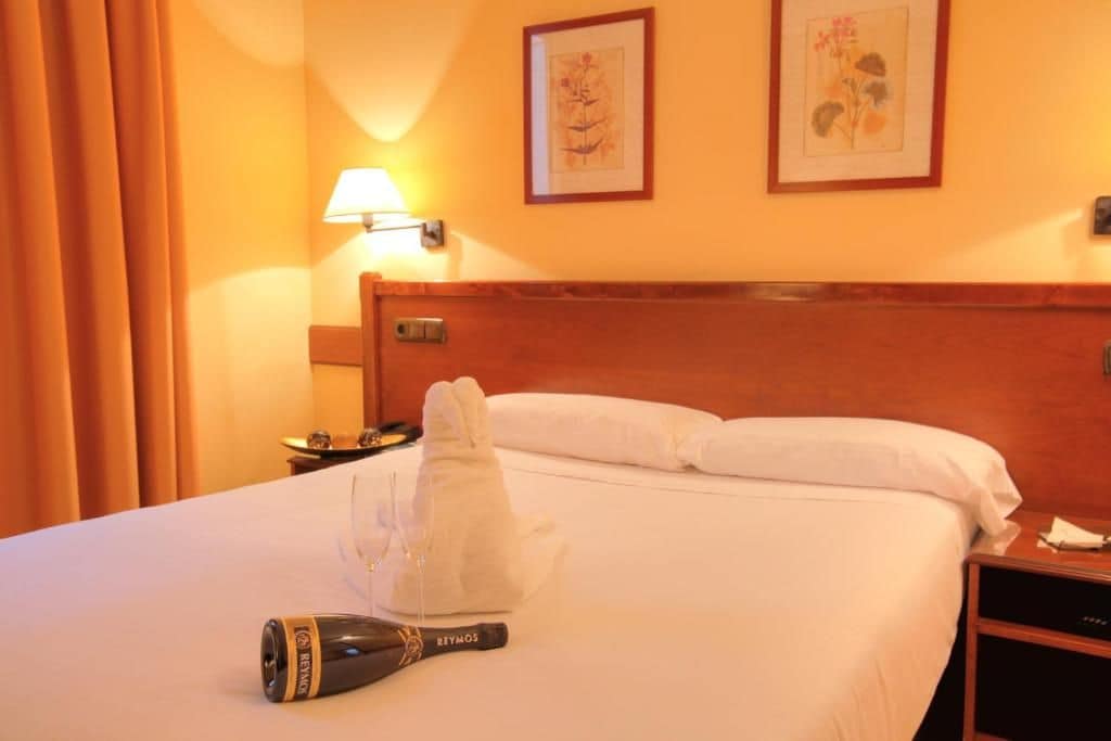 Une chambre d’hôtel avec une tête de lit en bois, un lit recouvert de draps blancs, une bouteille de vin Reymos et deux reproductions encadrées de motifs botaniques accrochées au mur.