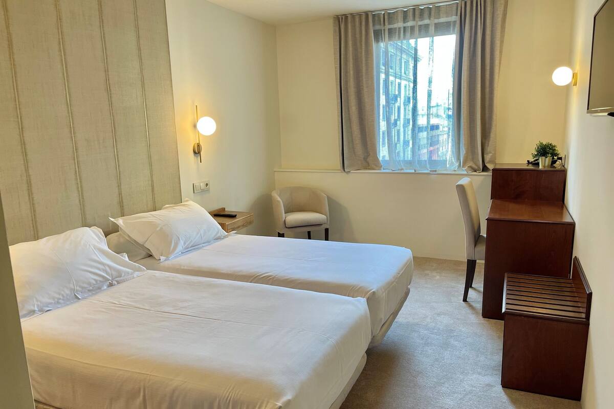 A hotel room with two beds, a desk, a chair, a window with curtains, and a wall-mounted TV.