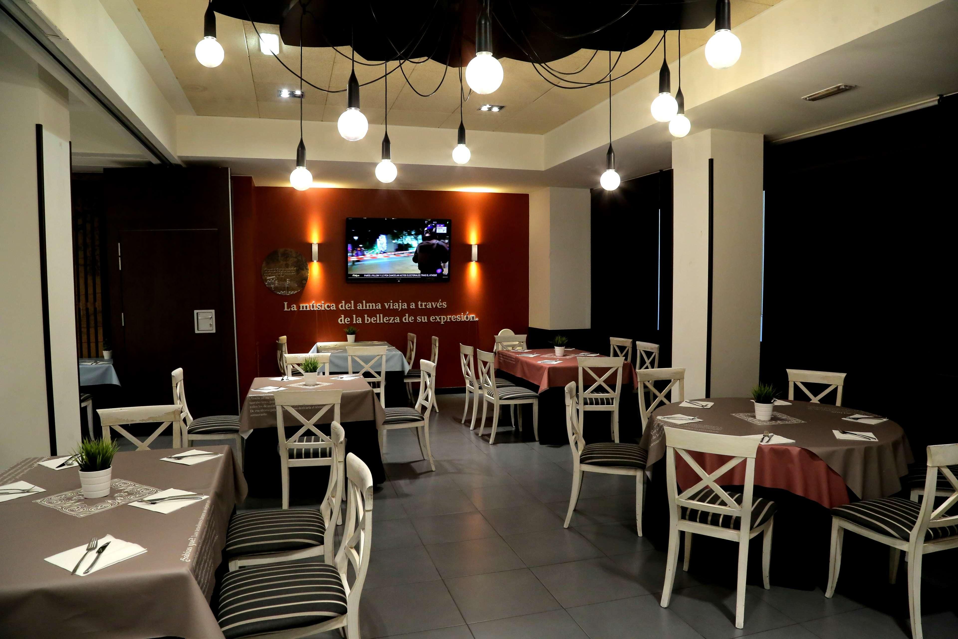 A restaurant with multiple tables and chairs, a television screen, and hanging light fixtures.