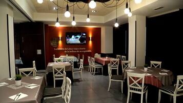 A restaurant with multiple tables and chairs, a television screen, and hanging light fixtures.