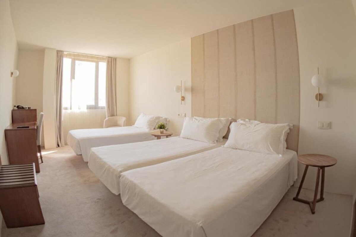 Habitación de hotel con dos camas, un escritorio, una silla, una mesita y una ventana con cortinas.