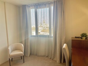 Ein Zimmer mit einem großen Fenster, das durch transparente Vorhänge verdeckt ist, einem beigefarbenen Sessel, einem Holztisch und einer Topfpflanze.