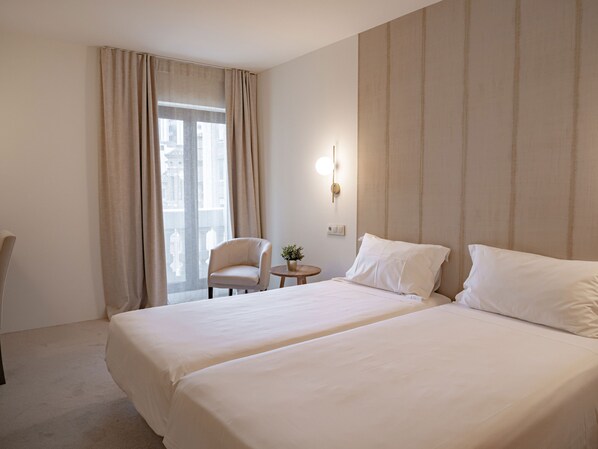 A hotel room with two beds, a chair, a small table, and a window with curtains.