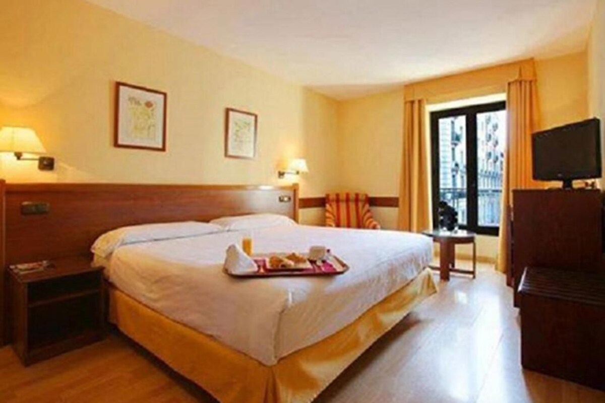 Una habitación de hotel con una cama grande, mesitas de noche, un televisor y una ventana con cortinas.