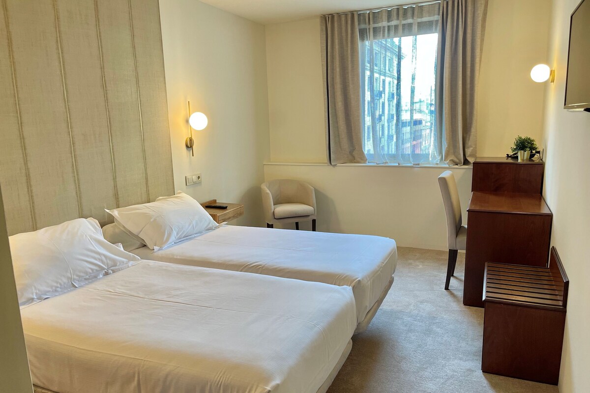 Habitación de hotel con dos camas, un escritorio, una silla, un ventanal con cortinas y un televisor montado en la pared.