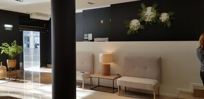 Eine Lobby mit Empfangsbereich, zwei Sitzgruppen, einer Wandpflanze und einer Tür mit der Beschriftung 'SALIDA'.