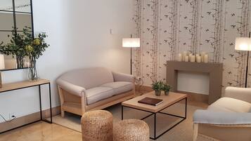 A living room with a beige sofa, a coffee table, wicker ottomans, a side table with a lamp, a mirror, and a fireplace with candles.
