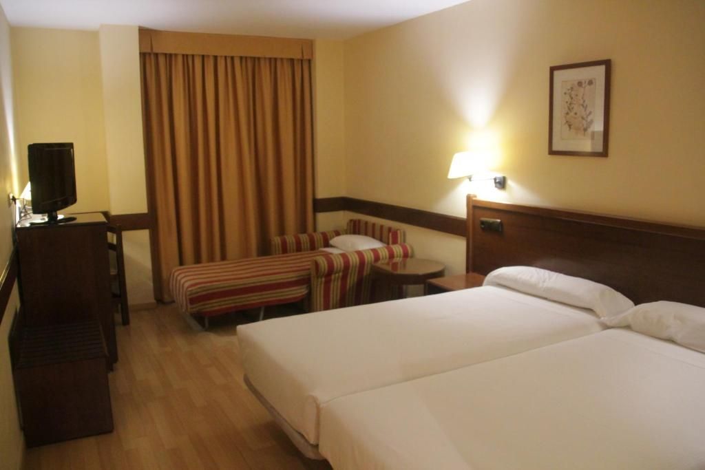 A hotel room with two beds, a desk with a television, a striped armchair, a small table, and a lamp.