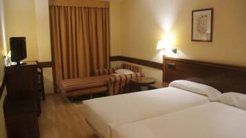 A hotel room with two beds, a desk with a television, a striped armchair, a small table, and a lamp.