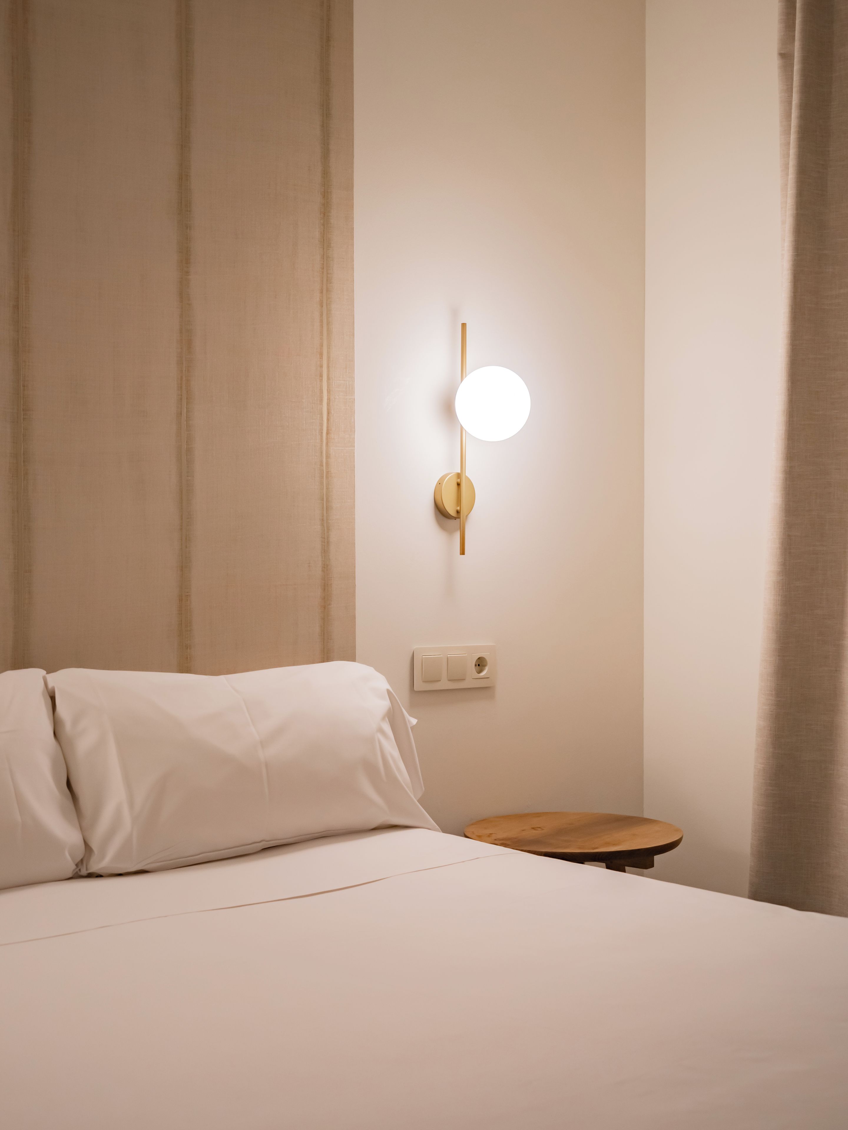 Hotellihuone, jossa on sänky, yöpöytä, seinään kiinnitetty valaisin ja pistorasia.