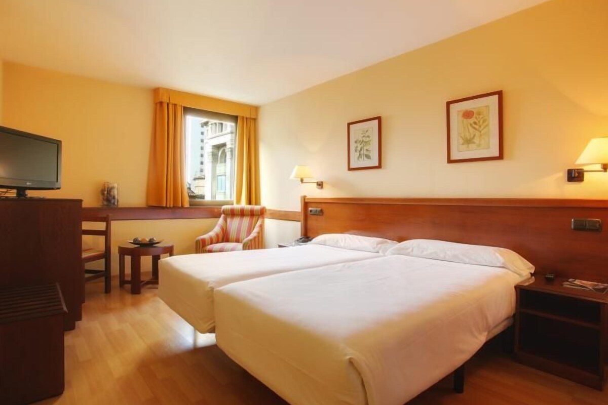 Habitación de hotel con dos camas, un escritorio con silla, televisor y obras de arte en las paredes.