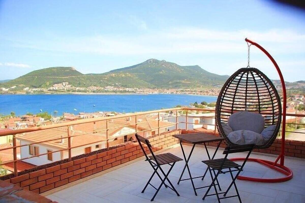 A balcony with a hanging chair, two chairs, and a table overlooking a coastal area with mountains in the background.