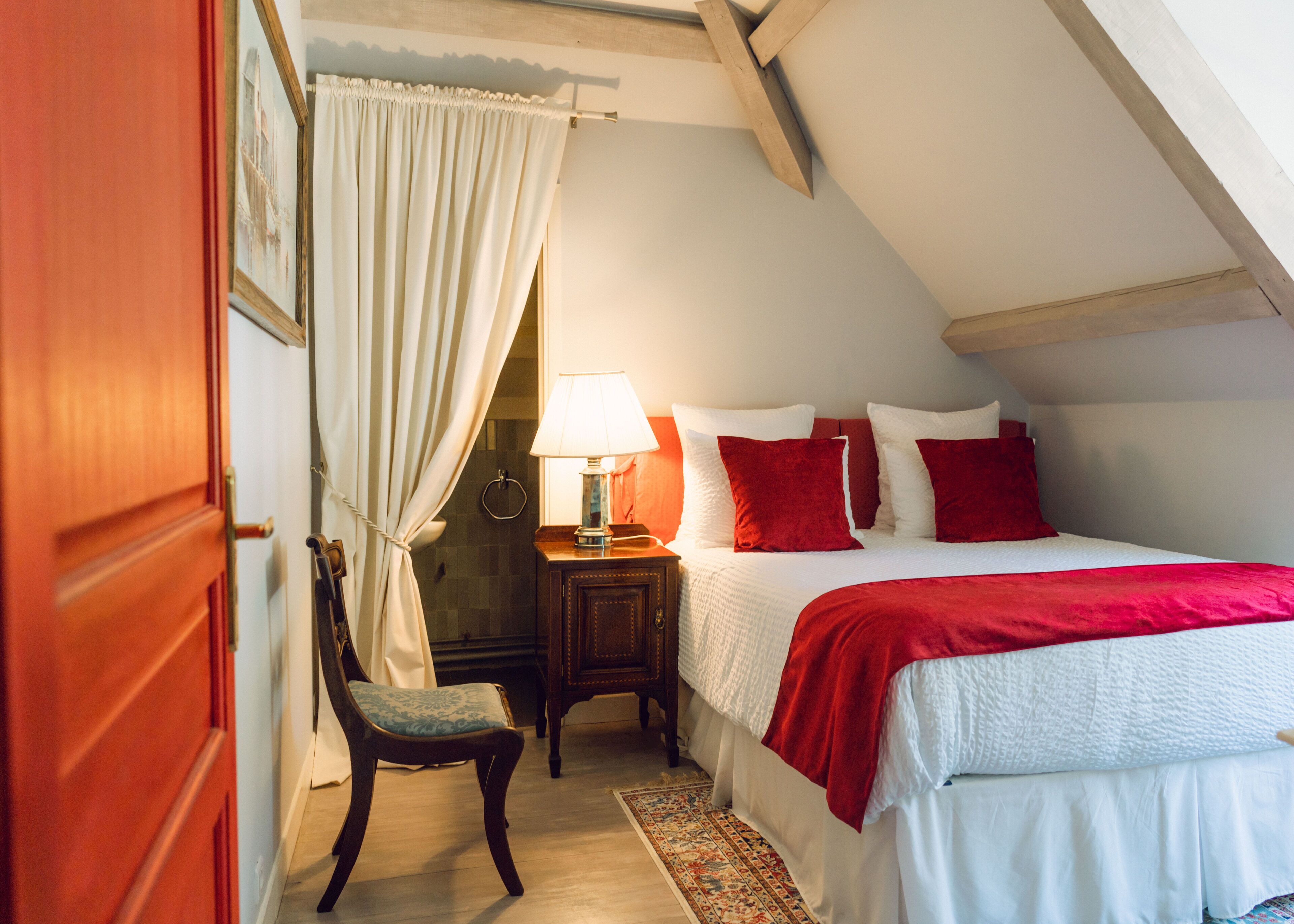 Un dormitorio con techo inclinado, cama con ropa de cama roja y blanca, mesita de noche con lámpara, una silla y un armario de madera.