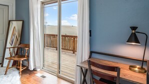 Ein Zimmer mit Holzboden, einem Holzstuhl, einem Schreibtisch mit Lampe und einer großen Schiebeglasdoor zu einem Balkon mit Holzgeländer.