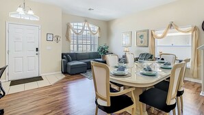A dining area with a round table, chairs, and a chandelier. There is a sofa and a window with curtains in the background. - Triton Pool/Spa  Home Near Disney With Spa & Resort Perks (Clermont)
